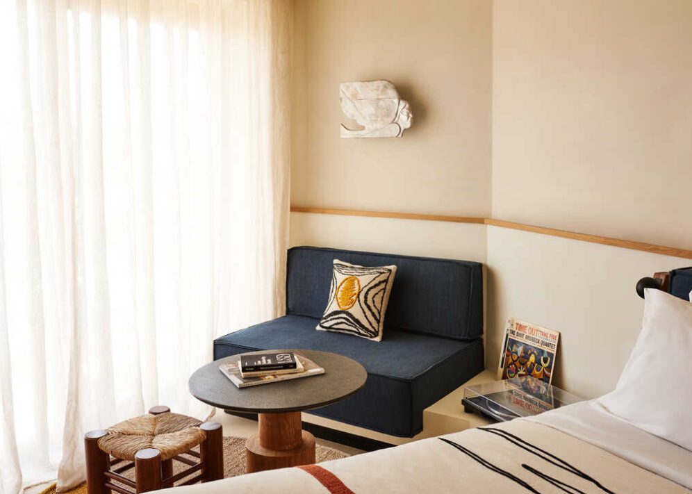 A blue chair and wooden stool sit next to a wooden and stone coffee table with reading material