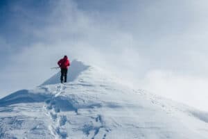 Person in a red jacket climbing a snow-covered mountain peak with ski poles, under a clear blue sky.