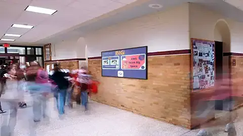 A large screen is mounted to a schools wall. Brick wall in the hallway with students walking by.