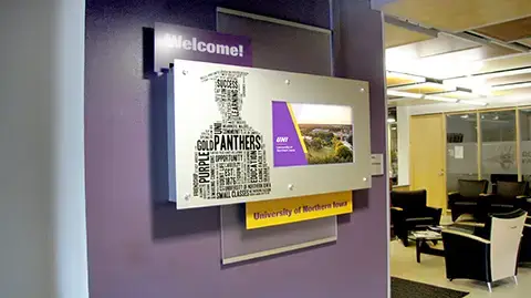 At the entry door of a university admissions office is a custom layered welcome display. The walls is painted in purple and the enclosure has graphis and the words welcome. The digital signage screen plays marketing messages to visitors