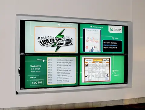 On a school wall an opening holds 4 screens. The screens are showing school digital signage content for events, menus, annoucements