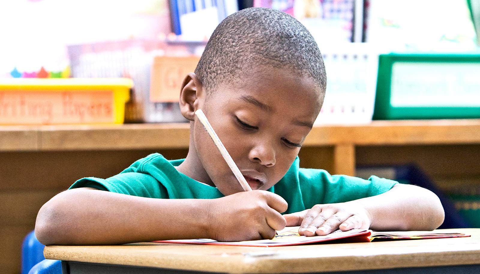 A young boy in the classroom writes into a notebook while at his desk.