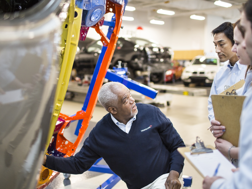 Plant manager kneels by machine equipment why explaining safety measures to inspectors with notepads. 