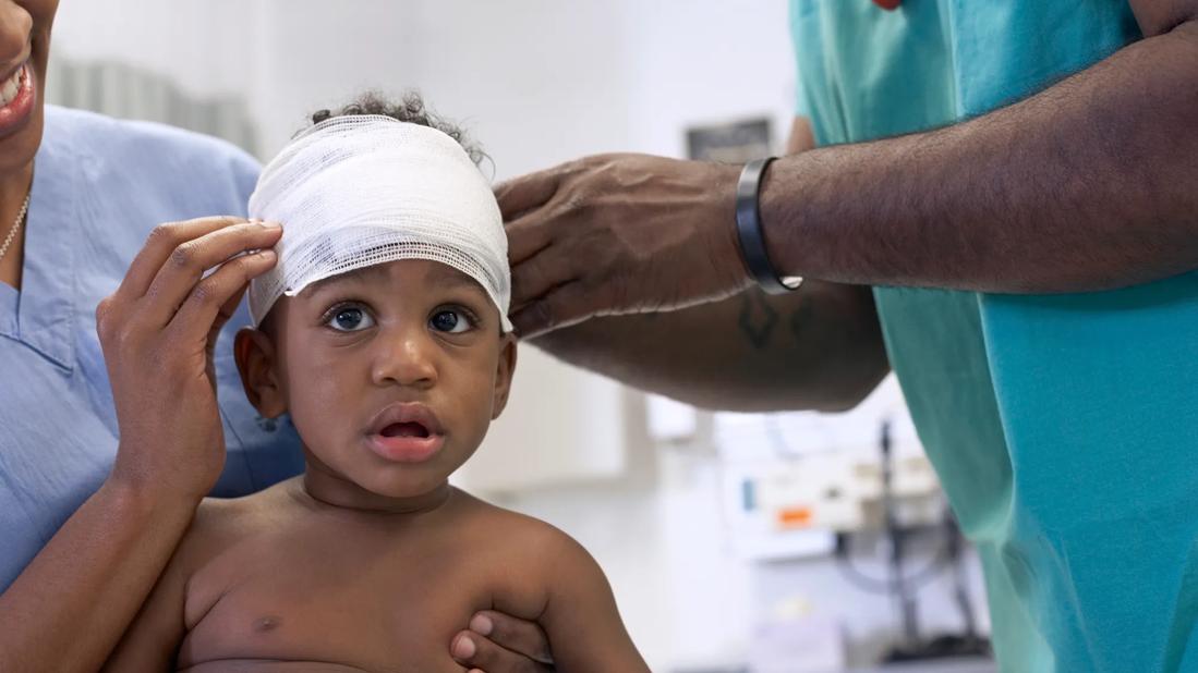 Toddler getting their head wrapped in bandage by two healthcare workers