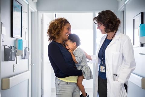 Smiling caregiver holding smiling toddler interacting with smiling healthcare provider in medical hallway