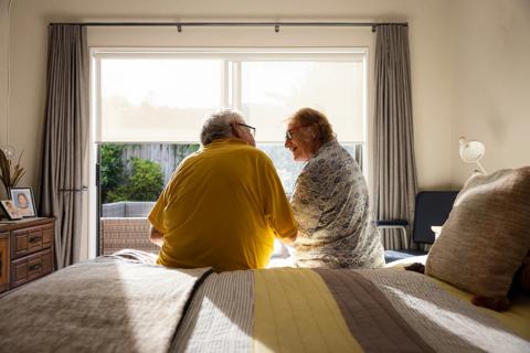 Elderly couple sitting on bed talking in bedroom, in early morning light