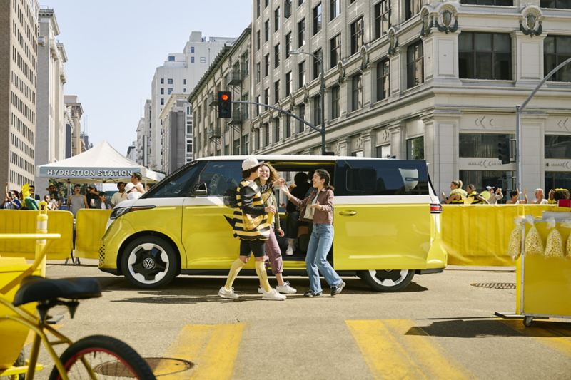 Profile view of an ID. Buzz shown in Pomelo Yellow Metallic and Candy White parked in downtown setting with event barriers and booths around the vehicle. There are people greeting each other outside the vehicle and one person is wearing a bumble bee costume.