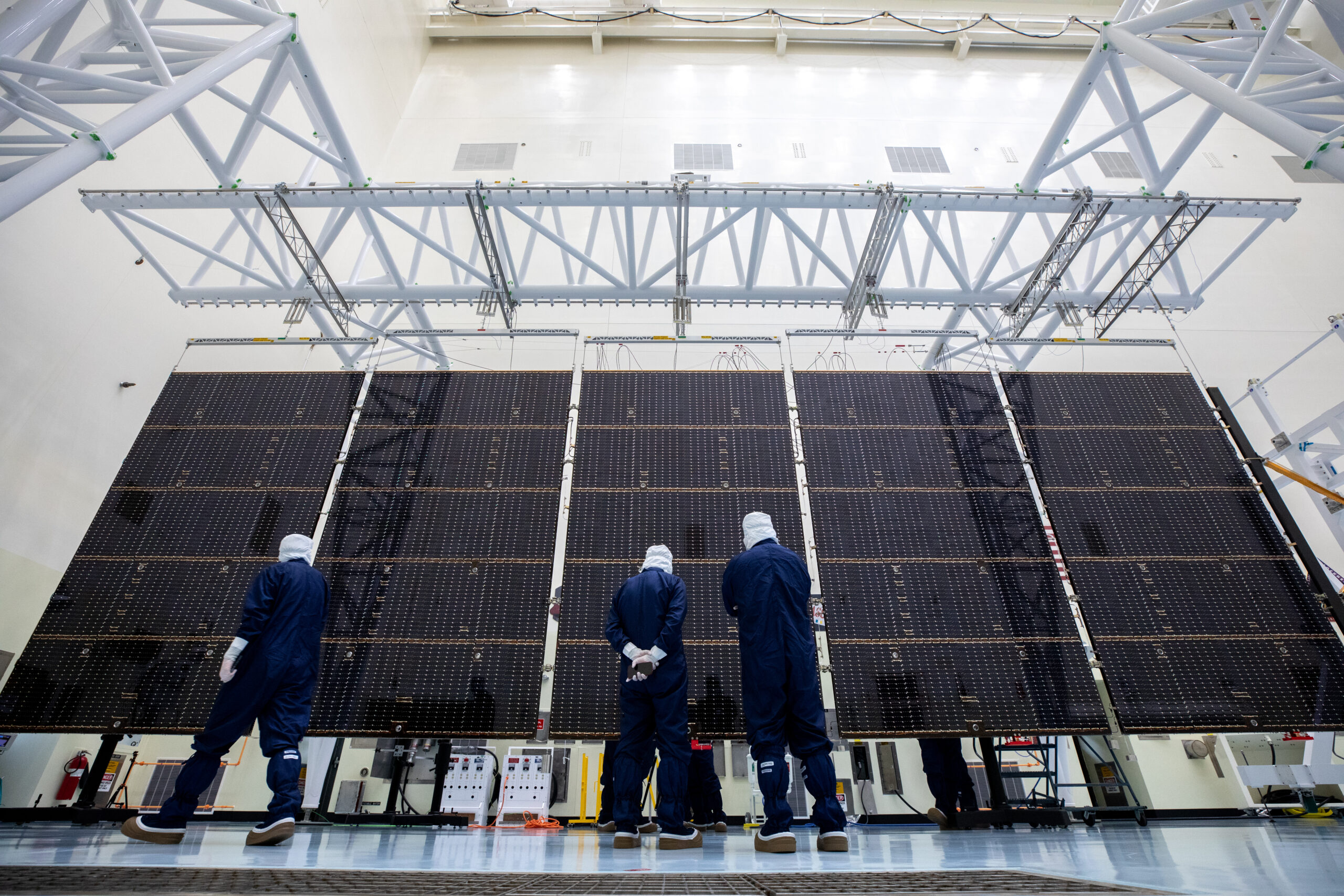 Three people in jumpsuits stand in front of a stretched out five-panel solar array inside of a building. 