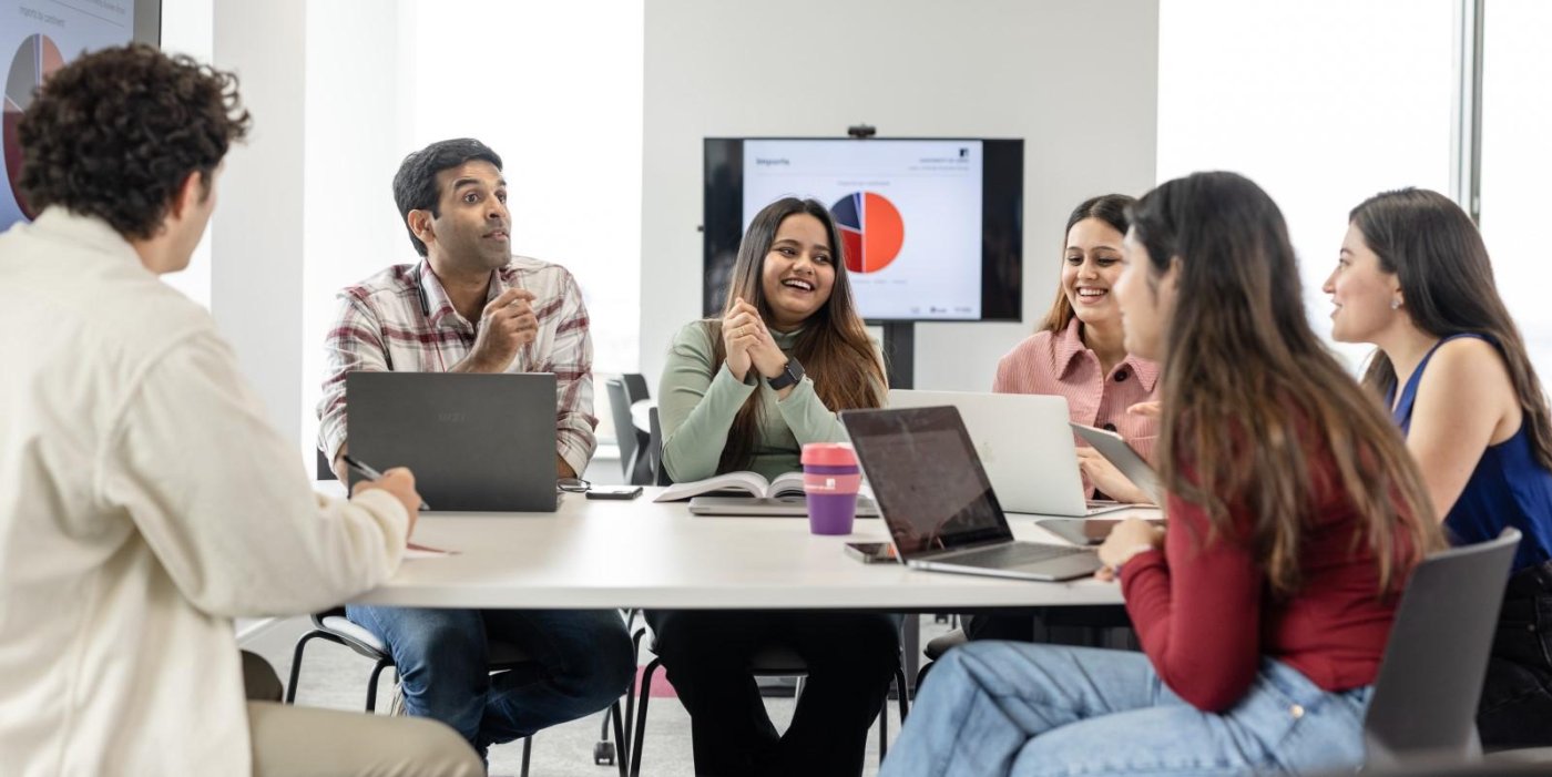 A group of Masters students interact around a table in a collaborative suite. There are laptops, books and cups on the table.