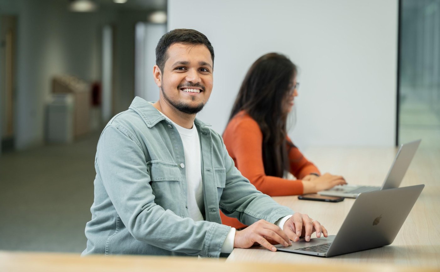 Student working at laptop smiles to camera. There is another student working in the background.