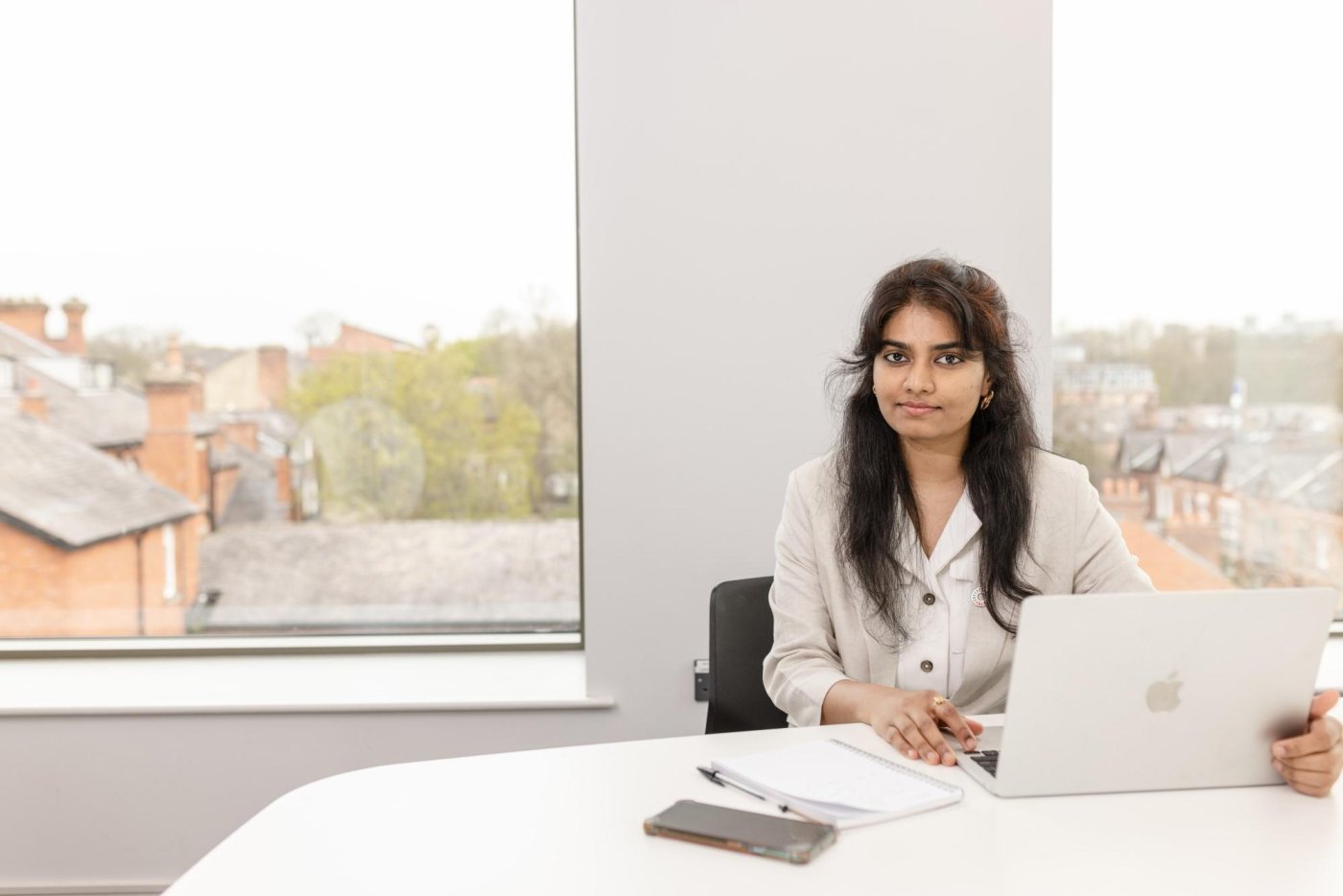 MBA student sitting with laptop and papers in room with large windows.