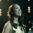 A woman in front of a microphone, looking to the right and smiling. She wears a gold vest and orange lights shine behind her.