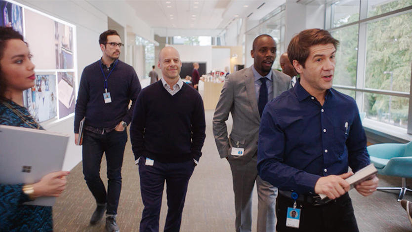 John Thompson from the SBC video is leading a group of five office workers down an office hallway while they carry on a conversation.