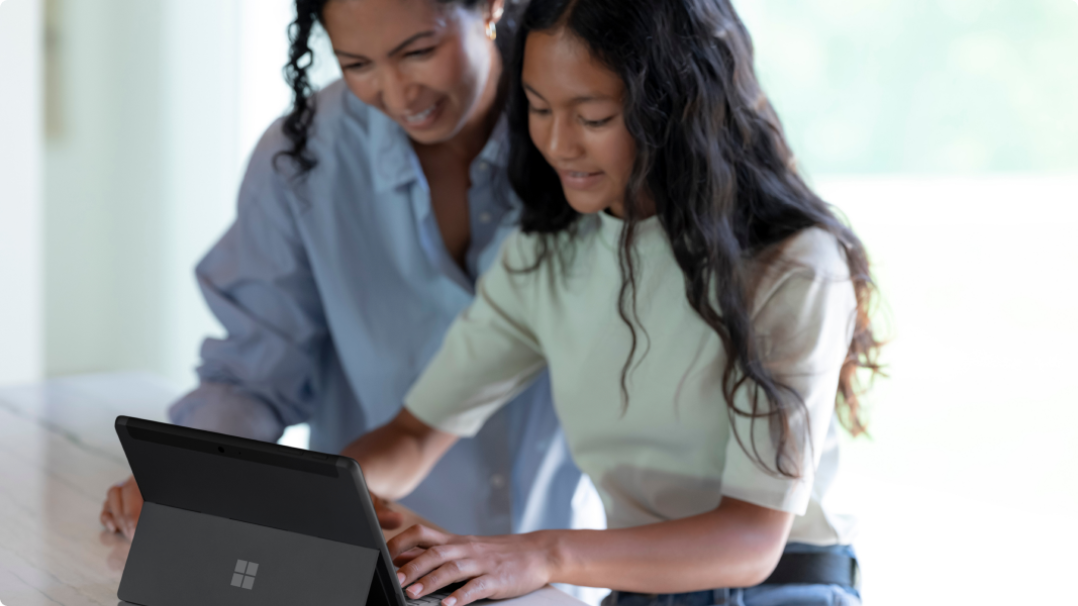 While one woman is typing on her Surface Pro computer, another woman watches over her shoulder