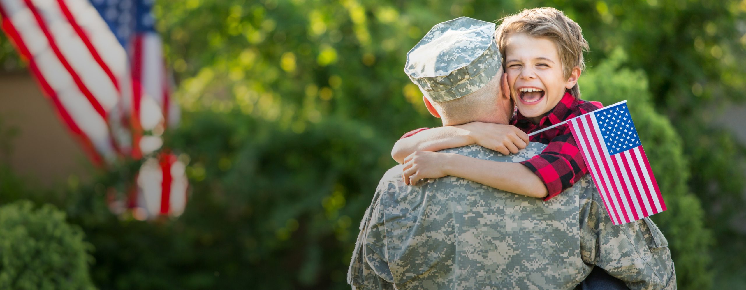 A smiling boy holding a small American flag hugging his father who is wearing military fatigues