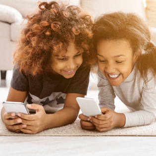 Two children lay on the ground while looking at their phones. 