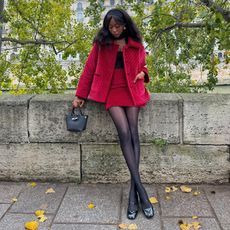 Emmanuelle is wearing a red velvet jacket, red velvet skirt, black top, black sheer tights, black ballet flats, black top-handle bag.