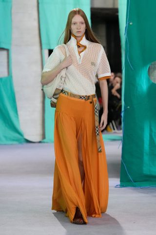 a model wears an orange and white polo shirt, an orange maxi skirt, burberry belt, and white shoulder bag.