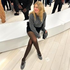 Danish fashion influencer Sidsel Alling sitting ona white bench at the Loewe fashion show wearing a gray polo sweater, black feather mini skirt, brown sheer tights, and studded Alaia Mary Jane flats