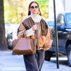 Katie Holmes wears a white turtleneck sweater, mixed color jacket, a cognac-color tote bag, gray wide-leg jeans, and black square-toe loafers.