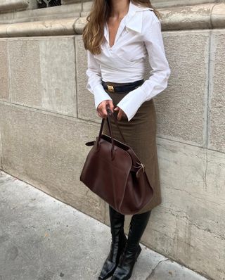 Woman wearing white button-down shirt, suede pencil skirt, black boots, and brown The Row Margaux tote bag.