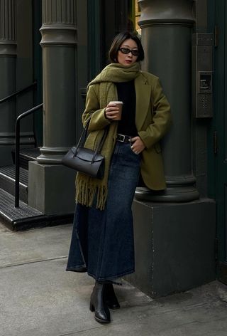 A denim skirt outfit is shown in a photo of a woman on the sidewalk wearing an oversized green blazer with a matching scarf, a black sweater, a black belt, a denim mini skirt, black boots, and a black shoulder bag