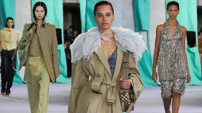 Models at the Burberry S/S 25 show. Model on the left wears a biege utilitarian jacket with gold pants. Model in the middle wears a beige trench coat with a fur collar and a shoulder saddle bag. Model on the right wears a halterneck sequin midi dress.