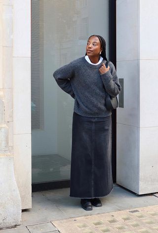 A denim skirt outfit is shown in a photo of a woman on the sidewalk wearing a white t-shirt under a gray sweater styled with a gray denim maxi skirt, black boots, and a black shoulder bag