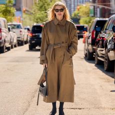 Courtney wears a long beige trench coat, black shoes, black sunglasses, and a gray bag.