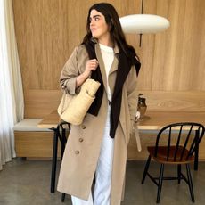 British fashion influencer Anna Howard poses in her modern London kitchen wearing a long trench coat, black sweater draped over her shoulders, a white t-shirt, belt, white pants, and a straw tote bag