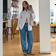 fashion influencer Sasha Mei poses for a mirror selfie wearing a white button-down shirt, cream Bottega Veneta shoulder bag, black belt, relaxed jeans, and black flip flops