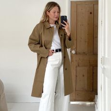 British influencer Alexis Foreman poses for a mirror selfie in a bright bedroom with white wood floors wearing a classic tan trench coat, white t-shirt, black belt, and white jeans