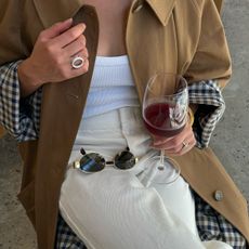 Sasha wears a trench coat, silver circle ring, white tank, ivory pants, and oval sunglasses.