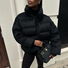 @nlmarilyn wearing a black puffer coat, leggings, tall boots, and an Hermès Birkin bag.