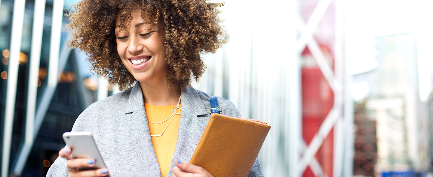 A person with curly hair smiling down at their smartphone and holding a tan notebook.