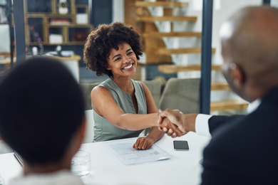 A person sitting across a desk from two interviewers, smiling and shaking hands with one of the interviewers.