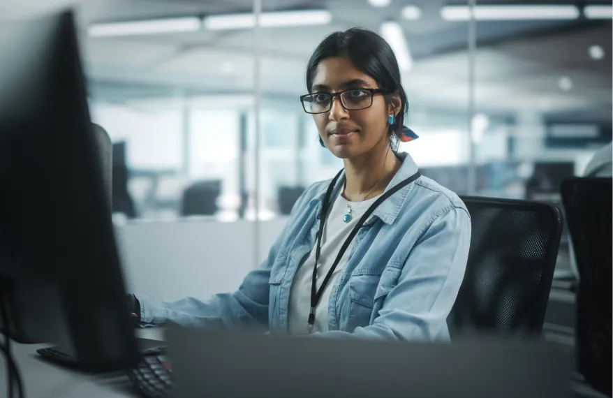 A woman wearing glasses and a blue jacket is sitting in front of a computer.