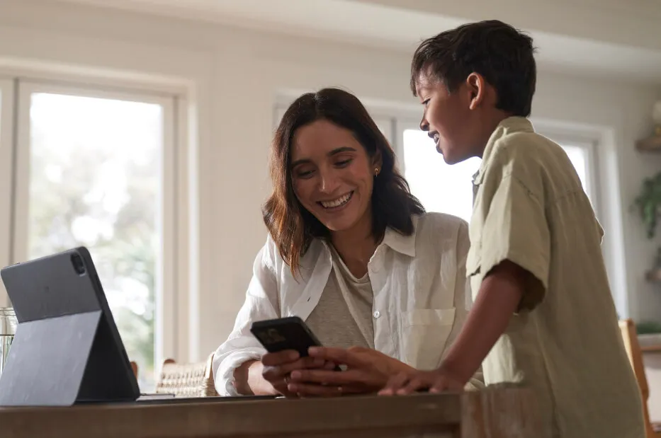 A woman sitting near a table and a young boy standing close to her, both smiling and looking a mobile phone.