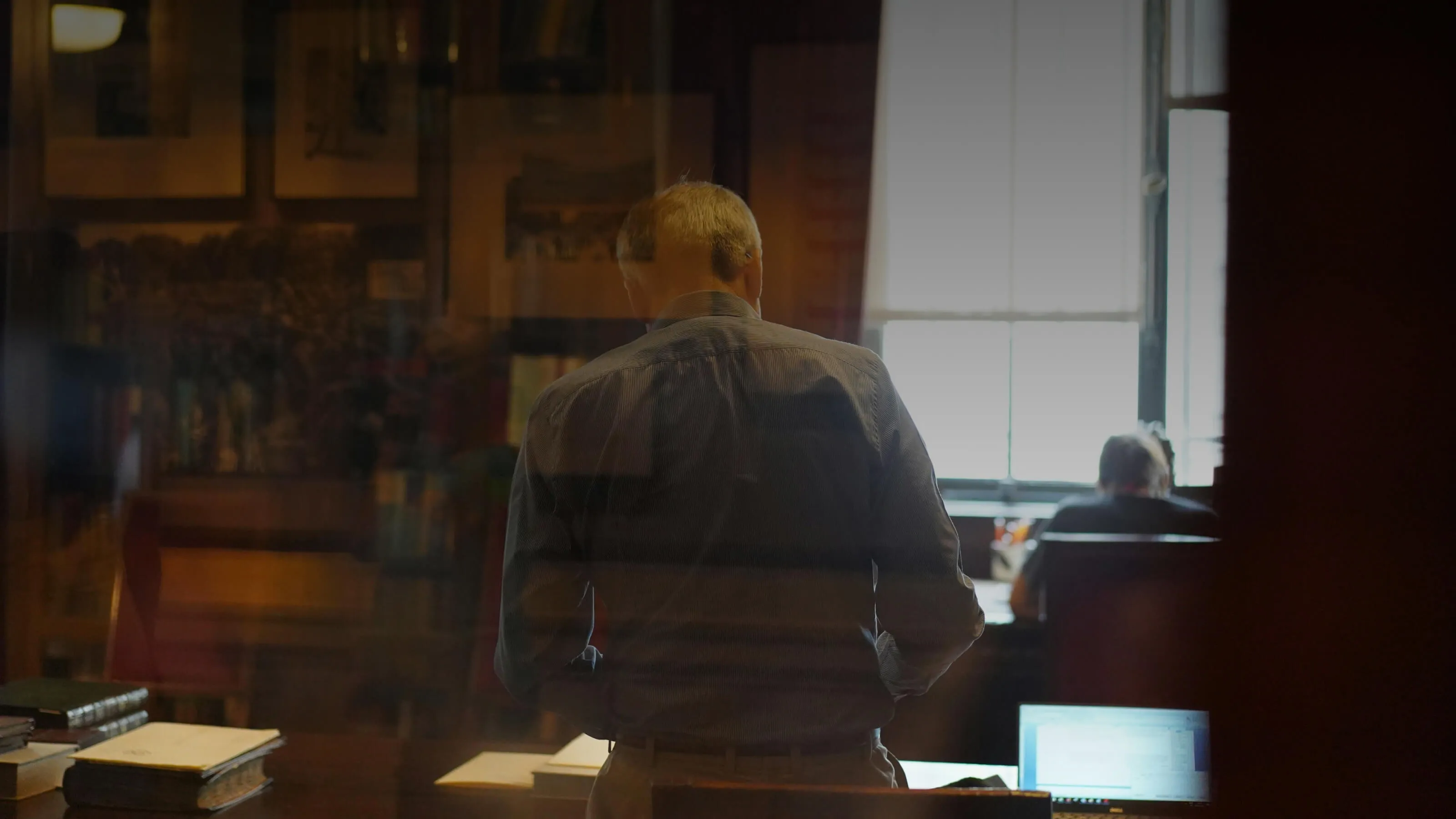 Man standing with his back to the camera in a dimly lit office or study, with books and papers on a desk, and another person seated at a desk near a window.