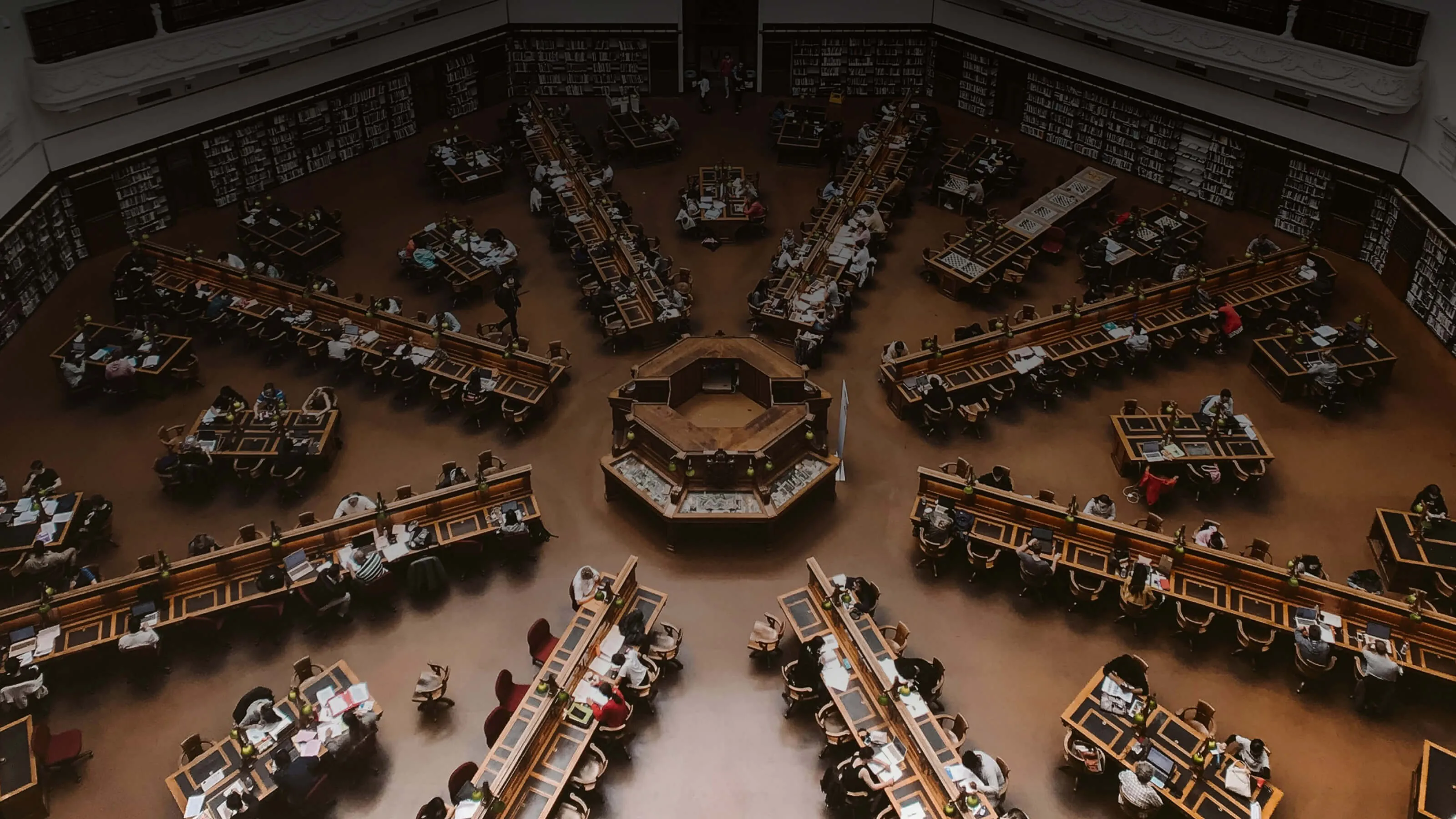 Large circular library reading room with long wooden tables arranged in a radial pattern, filled with people studying, and bookshelves lining the walls.