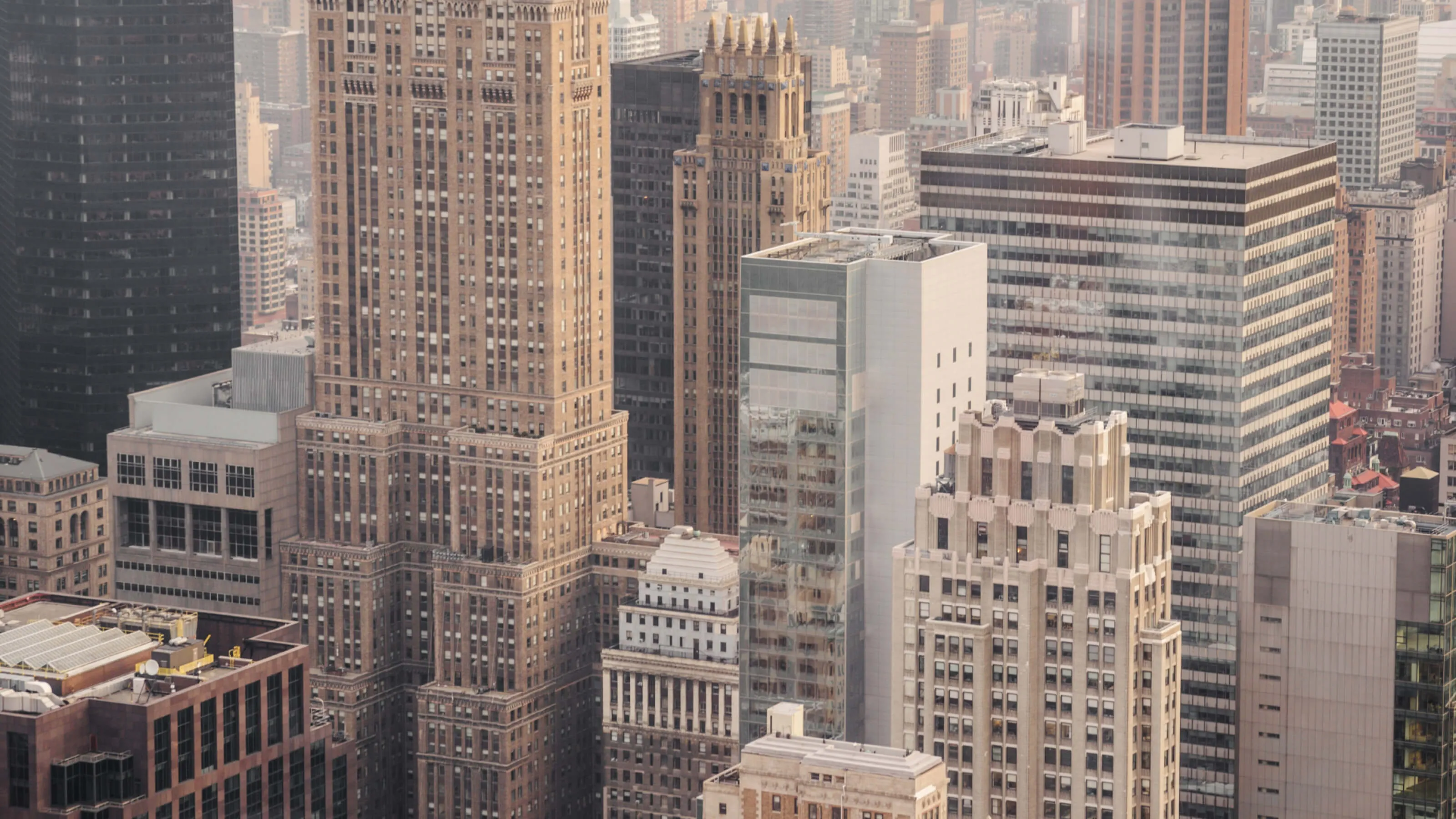 Cityscape with densely packed skyscrapers of varying architectural styles, including older brick buildings and modern glass structures, under soft, warm lighting.