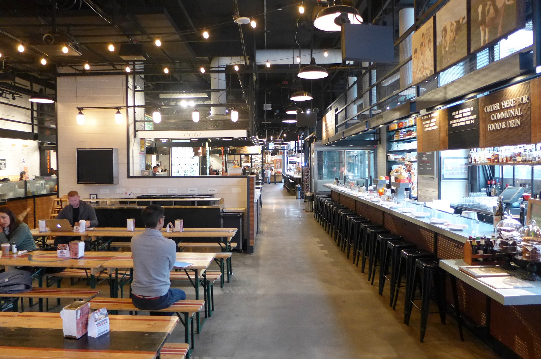 A person sits at a long wooden picnic bench located inside Gotham West Market.