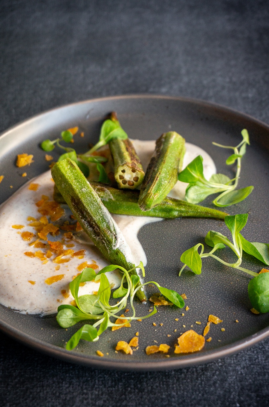 Slightly browned, large pieces of okra sit on a slate plate in a pool of a thin white sauce, garnished with greens and yellow crumbs.