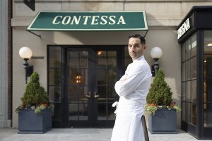 A chef stands in front of a restaurant with an awning that reads Contessa, his arms crossed, unsmiling.