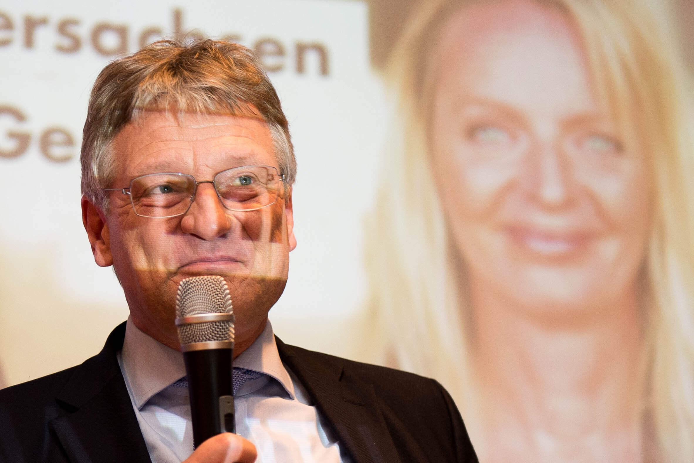 Jörg Meuthen steht auf einer Bühne und hält ein Mikrophon. Im Hintergrund ist ein Bild von Dana Guth groß an die Wand projeziert.