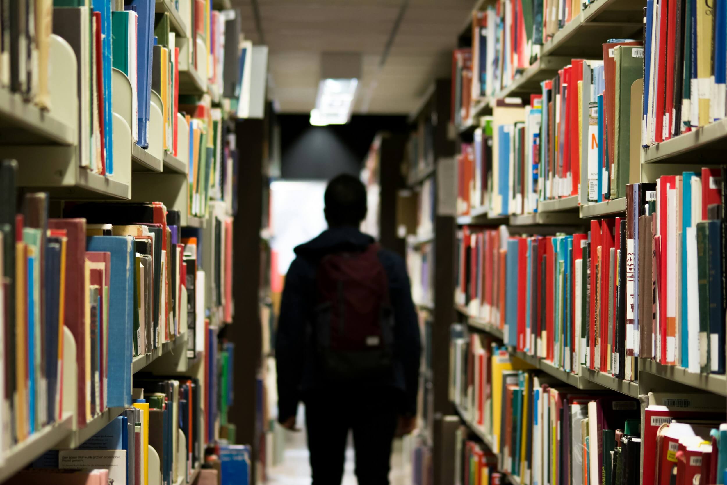 Ein Kind mit Schulranzen steht zwischen Bücherregalen in einer Bibliothek. Foto: redd-f-9o8Y / unsplash.com