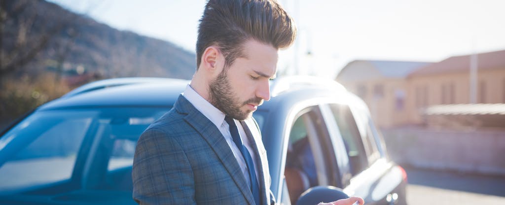 Man leaning against his car on a sunny day, reading on his phone about auto liability insurance