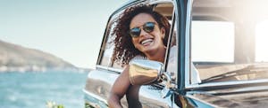 Woman sitting in her car, leaning out of the passenger side window on a sunny day, smiling