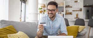 A smiling man wearing glasses holds a credit card while using a laptop.