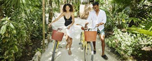 A smiling couple walk their bicycles along a sandy path surrounded by palms.